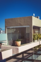A modern rooftop terrace is part of a square, minimalist building with a flat roof. The exterior walls are painted in muted earth tones. Large glass windows provide a view into the interior where a small table with chairs and a counter can be seen. The terrace features potted plants, including a large round planter with lush greenery, and several rectangular planters with taller plants. The sky is clear and deep blue.