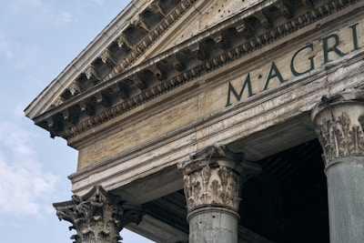 An angled view of an ancient Roman building showcasing intricate Corinthian columns and detailed carvings. The visible facade includes the partially seen inscription 'M·AGRIPPA'. The stone structure appears weathered, hinting at its historical significance. The sky in the background is partly cloudy.