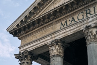 An angled view of an ancient Roman building showcasing intricate Corinthian columns and detailed carvings. The visible facade includes the partially seen inscription 'M·AGRIPPA'. The stone structure appears weathered, hinting at its historical significance. The sky in the background is partly cloudy.