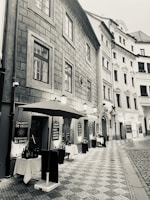 A narrow cobblestone street flanked by tall, historic buildings. On one side, an outdoor restaurant setting is visible with tables covered in white cloth, shaded by a large umbrella. Various menus are displayed on boards outside the restaurant, suggesting an Italian cuisine with options like spaghetti, tortellini, and desserts. The aesthetic is enhanced by classic street lamps and the architectural details of the buildings.