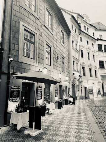A narrow cobblestone street flanked by tall, historic buildings. On one side, an outdoor restaurant setting is visible with tables covered in white cloth, shaded by a large umbrella. Various menus are displayed on boards outside the restaurant, suggesting an Italian cuisine with options like spaghetti, tortellini, and desserts. The aesthetic is enhanced by classic street lamps and the architectural details of the buildings.
