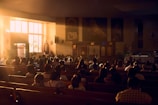 a group of people sitting in a room with a stage and a large screen