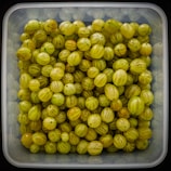 A collection of light green gooseberries fills a transparent square container. The berries are tightly packed, showcasing their translucent skin with faint stripes and small, dark specks at the ends.