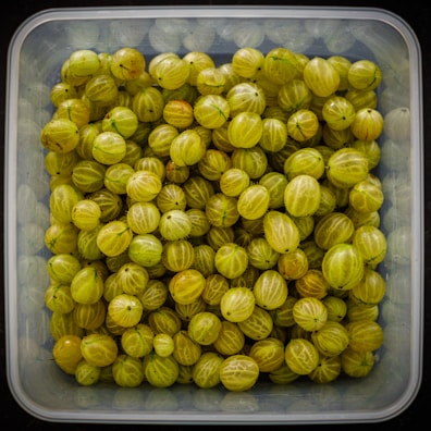 A collection of light green gooseberries fills a transparent square container. The berries are tightly packed, showcasing their translucent skin with faint stripes and small, dark specks at the ends.
