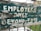 A weathered and peeling sign with the text 'EMPLOYEES ONLY BEYOND THIS' painted in white on a green background, mounted on dilapidated wood. The sign is outdoors, positioned in front of a grassy area and industrial buildings.