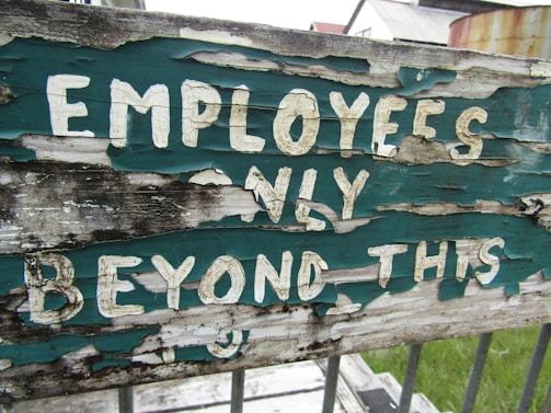 A weathered and peeling sign with the text 'EMPLOYEES ONLY BEYOND THIS' painted in white on a green background, mounted on dilapidated wood. The sign is outdoors, positioned in front of a grassy area and industrial buildings.
