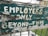 A weathered and peeling sign with the text 'EMPLOYEES ONLY BEYOND THIS' painted in white on a green background, mounted on dilapidated wood. The sign is outdoors, positioned in front of a grassy area and industrial buildings.