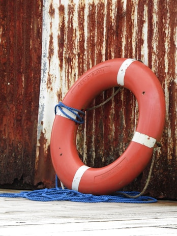A red lifebuoy with white stripes leaning against a rusted, distressed metal wall. It is resting on a wooden surface and is accompanied by a coiled blue rope.