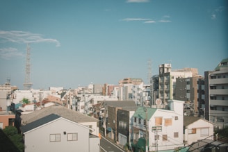 An urban landscape featuring a variety of buildings under a clear blue sky. Structures include residential and commercial buildings, some with modern designs and others with traditional rooftops. Electric poles and wires are visible, contributing to the urban atmosphere.