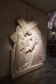 A detailed stone relief sculpture depicting a biblical scene with two figures, one carrying a cross. The figures are intricately carved, with flowing robes and expressions that suggest a moment of interaction or compassion. The relief is mounted on a wall in a dimly lit church interior, with wooden pews visible in the background.