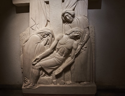 A detailed stone relief sculpture depicting three figures in a somber scene. The central figure appears lifeless and is supported by two others, one on each side. The figure on the left is cloaked and appears to be grieving, while the figure on the right has a mournful, tender expression. The background features draped cloth and a cross, adding to the religious and emotional context of the artwork.