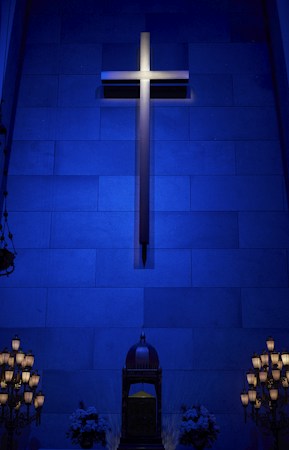 A large, illuminated cross is mounted on a blue-tinted wall made of rectangular stone tiles. Below the cross, there is a decorative altar flanked by arrangements of flowers. Elegant light fixtures with multiple bulbs frame the scene on either side, enhancing the atmospheric lighting.
