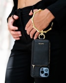 A person is holding a sleek black phone case with a gold metallic wristlet attached. The focus is on the elegant accessory, which contrasts against a black outfit. The person's hands are gently positioned near the waist, adding to the sophisticated appearance.