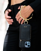 A person is holding a sleek black phone case with a gold metallic wristlet attached. The focus is on the elegant accessory, which contrasts against a black outfit. The person's hands are gently positioned near the waist, adding to the sophisticated appearance.