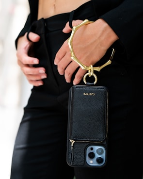 A person is holding a sleek black phone case with a gold metallic wristlet attached. The focus is on the elegant accessory, which contrasts against a black outfit. The person's hands are gently positioned near the waist, adding to the sophisticated appearance.