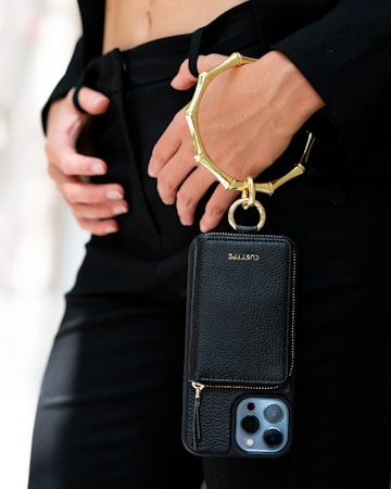 A person is holding a sleek black phone case with a gold metallic wristlet attached. The focus is on the elegant accessory, which contrasts against a black outfit. The person's hands are gently positioned near the waist, adding to the sophisticated appearance.