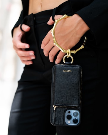 A person is holding a sleek black phone case with a gold metallic wristlet attached. The focus is on the elegant accessory, which contrasts against a black outfit. The person's hands are gently positioned near the waist, adding to the sophisticated appearance.