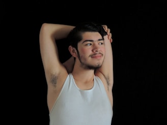 A person wearing a white tank top with a relaxed posture, arms raised behind the head, against a black background. The individual has facial hair and a slight smile, conveying a calm and confident demeanor.