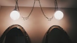 Two ornate mirrors with carved wooden frames are mounted on a beige wall. Above them, two spherical light fixtures hang from chains, casting a warm glow.