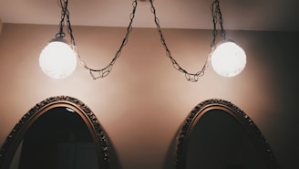 Two ornate mirrors with carved wooden frames are mounted on a beige wall. Above them, two spherical light fixtures hang from chains, casting a warm glow.