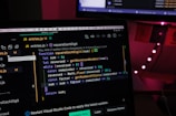 A laptop screen displaying a JavaScript code editor, focusing on a function called squareEachDigit. The environment is dimly lit with the background showing faint red lights and a window with partially visible curtains.