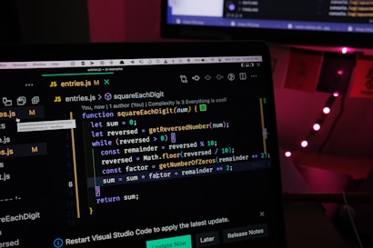 A laptop screen displaying a JavaScript code editor, focusing on a function called squareEachDigit. The environment is dimly lit with the background showing faint red lights and a window with partially visible curtains.