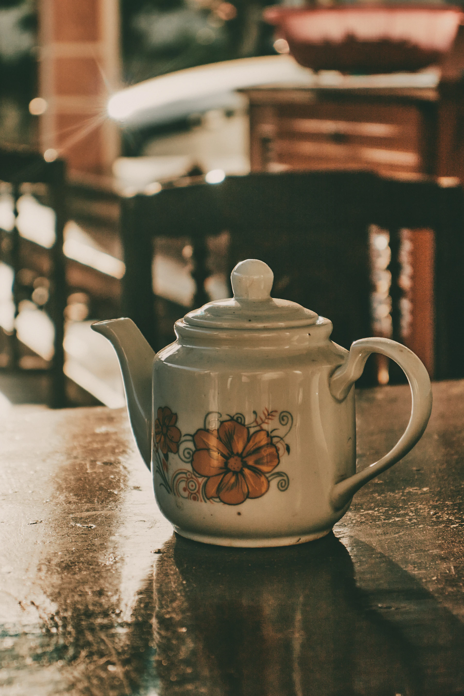 A white teapot printed with an orange flower, standing on a wet table in low sunlight