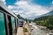 A scenic mountain road with buses traveling along a river, surrounded by lush green forest and distant snow-capped mountains under a partly cloudy sky.