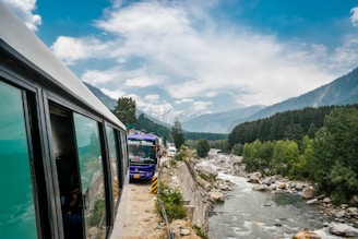 A scenic mountain road with buses traveling along a river, surrounded by lush green forest and distant snow-capped mountains under a partly cloudy sky.