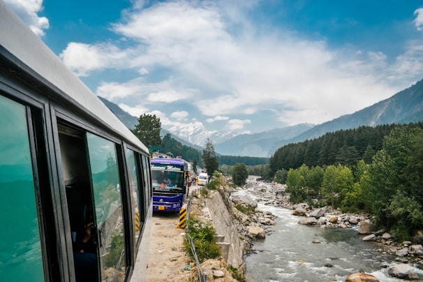 A scenic mountain road with buses traveling along a river, surrounded by lush green forest and distant snow-capped mountains under a partly cloudy sky.