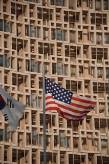 Two flags, the American flag and another with red, blue, and black elements, are prominently displayed against the backdrop of a beige building with a grid-like pattern of windows.