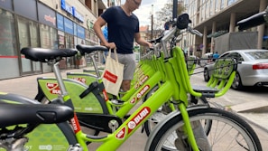 A person is standing next to a row of bright green rental bicycles in an urban setting. The bicycles are branded with 'MOL Bubi' and are aligned neatly on a sidewalk. The background includes several storefronts and a parked car, suggesting a busy city street.