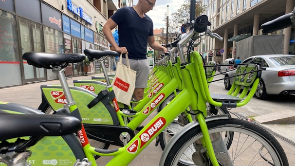 A person is standing next to a row of bright green rental bicycles in an urban setting. The bicycles are branded with 'MOL Bubi' and are aligned neatly on a sidewalk. The background includes several storefronts and a parked car, suggesting a busy city street.