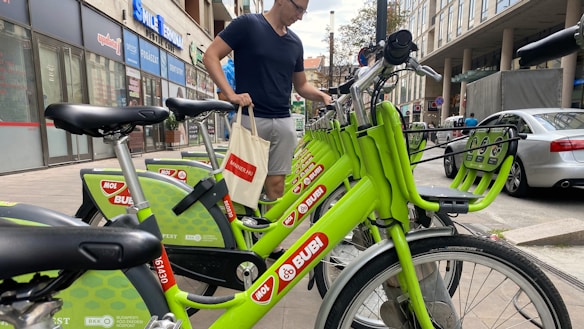 A person is standing next to a row of bright green rental bicycles in an urban setting. The bicycles are branded with 'MOL Bubi' and are aligned neatly on a sidewalk. The background includes several storefronts and a parked car, suggesting a busy city street.