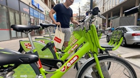 A person is standing next to a row of bright green rental bicycles in an urban setting. The bicycles are branded with 'MOL Bubi' and are aligned neatly on a sidewalk. The background includes several storefronts and a parked car, suggesting a busy city street.