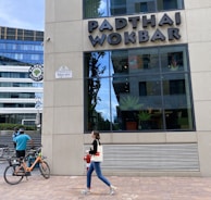 A modern building exterior features the sign 'PADTHAI WOKBAR'. A person walks on the sidewalk carrying a bag and drink. Bicycles are parked nearby, with a cyclist standing next to them.