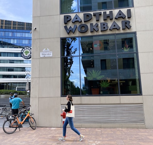 A modern building exterior features the sign 'PADTHAI WOKBAR'. A person walks on the sidewalk carrying a bag and drink. Bicycles are parked nearby, with a cyclist standing next to them.