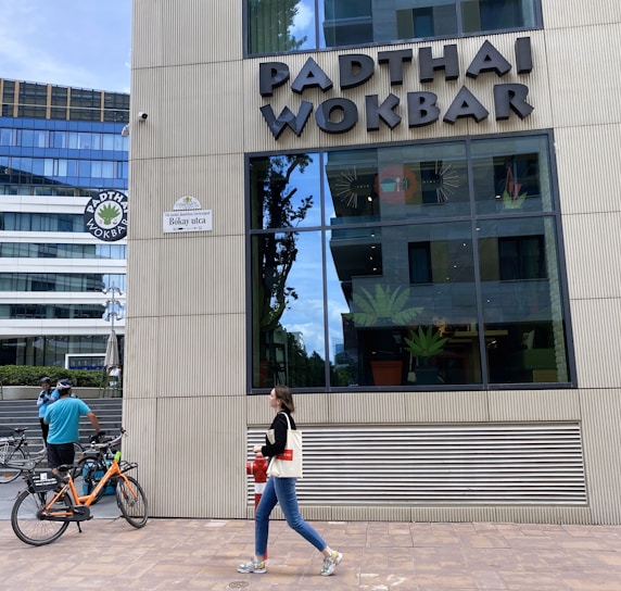A modern building exterior features the sign 'PADTHAI WOKBAR'. A person walks on the sidewalk carrying a bag and drink. Bicycles are parked nearby, with a cyclist standing next to them.
