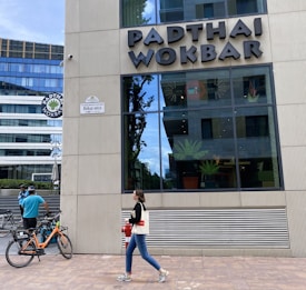 A modern building exterior features the sign 'PADTHAI WOKBAR'. A person walks on the sidewalk carrying a bag and drink. Bicycles are parked nearby, with a cyclist standing next to them.