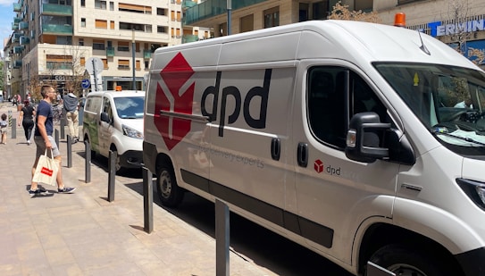 A white delivery van with the logo 'dpd' is parked along a city street. Several people walk by, including a man carrying a tote bag with a red design. Other vehicles are also parked nearby, and modern apartment buildings line the street.