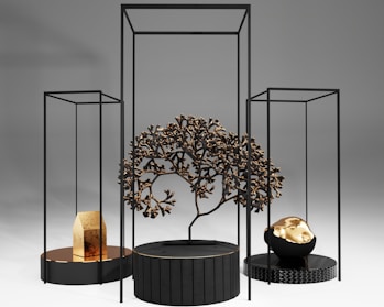 A modern art installation featuring geometric black frames and circular platforms. A central black platform holds a metallic tree with intricate branches. To the left, a smaller platform displays a gold object resembling a house. On the right, another platform supports a black and gold rock-like sculpture. The background is a smooth gradient of light to dark gray.