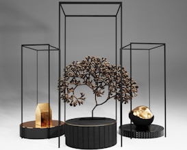 A modern art installation featuring geometric black frames and circular platforms. A central black platform holds a metallic tree with intricate branches. To the left, a smaller platform displays a gold object resembling a house. On the right, another platform supports a black and gold rock-like sculpture. The background is a smooth gradient of light to dark gray.
