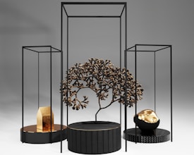 A modern art installation featuring geometric black frames and circular platforms. A central black platform holds a metallic tree with intricate branches. To the left, a smaller platform displays a gold object resembling a house. On the right, another platform supports a black and gold rock-like sculpture. The background is a smooth gradient of light to dark gray.