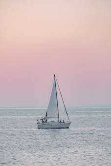 A sailboat glides smoothly across calm waters during a serene sunset. The sky is painted in soft pastel shades of pink and peach, contrasting with the gentle ripples of the blue-gray sea. Several people are visible on the boat, suggesting a leisurely cruise.