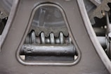Close-up view of a mechanical gear system, featuring large metal components with visible gears and axles. The metal surfaces are painted in a matte gray color, with signs of wear and some accumulated grease at the joints.