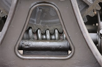 Close-up view of a mechanical gear system, featuring large metal components with visible gears and axles. The metal surfaces are painted in a matte gray color, with signs of wear and some accumulated grease at the joints.