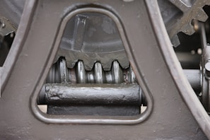 Close-up view of a mechanical gear system, featuring large metal components with visible gears and axles. The metal surfaces are painted in a matte gray color, with signs of wear and some accumulated grease at the joints.