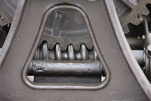 Close-up view of a mechanical gear system, featuring large metal components with visible gears and axles. The metal surfaces are painted in a matte gray color, with signs of wear and some accumulated grease at the joints.