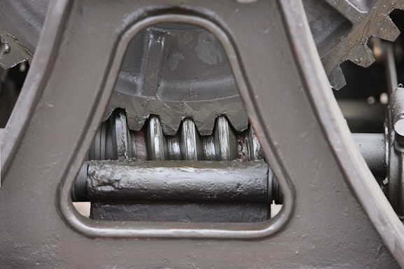 Close-up view of a mechanical gear system, featuring large metal components with visible gears and axles. The metal surfaces are painted in a matte gray color, with signs of wear and some accumulated grease at the joints.