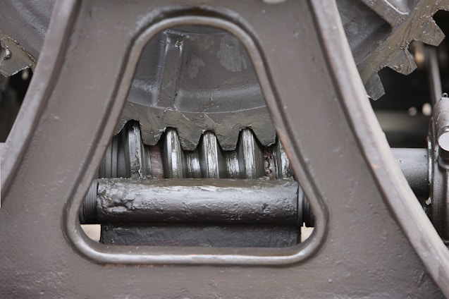 Close-up view of a mechanical gear system, featuring large metal components with visible gears and axles. The metal surfaces are painted in a matte gray color, with signs of wear and some accumulated grease at the joints.